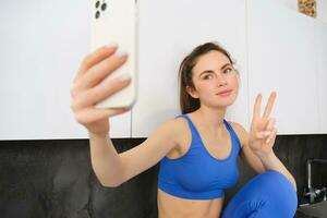 passen und gesund jung Sport Frau, zeigen Frieden Zeichen, nehmen Selfie im Aktivkleidung, Fitness Blogger Herstellung Fotos zum Sozial Medien im ihr Activewear