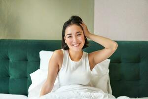 süß Koreanisch Mädchen im Weiß Panzer Spitze, wacht auf oben im ihr Schlafzimmer, Lügen im Bett im Morgen, Dehnen Hände und lächelt foto