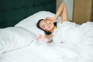 romantisch asiatisch Mädchen aufwachen oben im gemütlich Schlafzimmer, Lügen im Bett mit Weiß Blätter, Dehnen ihr Hand gegenüber leeren Kissen foto