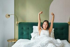Porträt von lächelnd glücklich asiatisch Mädchen, wacht auf oben Gefühl enthusiastisch, erstreckt sich ihr Hände hoch, genießt gut Morgen im ihr Schlafzimmer foto