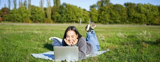 lächelnd asiatisch Mädchen Lügen im Park auf Gras, Aufpassen Film oder Video auf Laptop, suchen beim Bildschirm mit Interesse foto