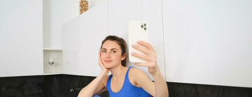 Porträt von Fitness Mädchen posieren zum Foto, nehmen Selfie auf Smartphone Anwendung, Sitzung im Küche, tragen Activewear foto