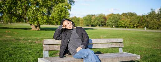 lächelnd jung asiatisch Frau im draussen Jacke, Sitzung auf Bank im Grün sonnig Park, ruhen allein, entspannend auf frisch Luft foto