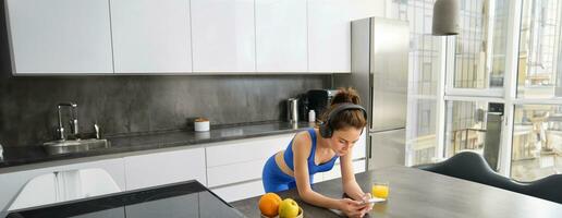 Porträt von Brünette Frau im Sportbekleidung, mit Smartphone, Stehen im Küche, Trinken frisch Orange Saft, Aufpassen trainieren Fitnessstudio Tutorials foto