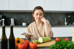 Frau Aufpassen Rezept auf Smartphone, Sitzung im das Küche mit Gemüse und Hacken Planke, vorbereiten Abendessen, gesund Salat, Kochen beim heim, tragen Bademantel foto