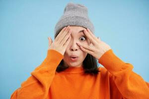 überrascht Koreanisch Mädchen im warm Hut, sieht aus erstaunt beim Kamera, drückt aus Interesse und Staunen, steht im Orange Sweatshirt Über Blau Hintergrund foto