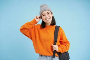 stilvoll jung asiatisch Frau im warm Hut, Gehen mit Rucksack, gehen irgendwo mit Tasche, Stehen Über Blau Studio Hintergrund foto