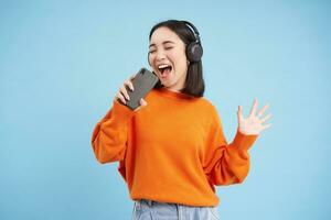 schön Koreanisch Mädchen Singen im Smartphone Mikrofon, hört zu Musik- im Kopfhörer, singt Karaoke auf Handy, Mobiltelefon Anwendung, steht Über Blau Hintergrund foto