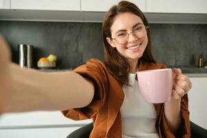 Porträt von schön jung Frau im Gläser, nehmen Selfie im Küche mit Tasse von Morgen Kaffee, Herstellung Fotos zum ihr Täglich Routine Lebensstil Blog