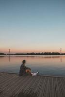 jung 25 Jahr alt braun Mann mit Plaid Hemd Sitzung auf das Ende von ein hölzern Seebrücke während Sonnenuntergang, Aufpassen das Ruhe Wasser Oberfläche im Almere Niederlande foto