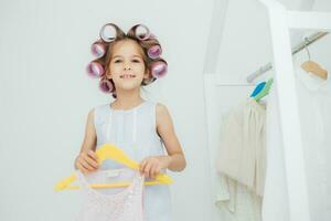 lächelnd Mädchen mit Haar Walzen wählen ein Kleid, spielerisch Ankleiden foto