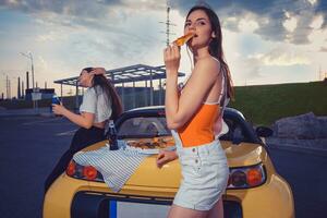 attraktiv Frauen im beiläufig Kleider Essen Pizza, genießen mit Kohlensäure trinken im Glas Flaschen während posieren in der Nähe von Gelb Auto auf Parkplatz viel. Kopieren Raum foto