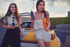 Mädchen im beiläufig Outfit sind Lachen und genießen Limonade im Glas Flaschen während gelehnt auf Kofferraum von Gelb Auto mit Pizza auf Es. schnell Lebensmittel. Kopieren Raum foto