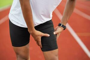 Sportler Sport Mann Läufer tragen Weiß Sportbekleidung zu Sitzung Gefühl Schmerzen im seine Bein oder Knie oder Schenkel nach üben auf ein Laufen Spur beim ein Stadion, Kopieren Raum. Läufer Sport Verletzung Konzept. foto