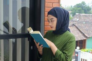 Porträt von attraktiv asiatisch Hijab Frau halten Buch im Balkon. Muslim Mädchen lesen ein Buch. Konzept von Alphabetisierung und Wissen foto