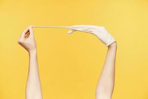 Studio Foto von jung Frau Hände posieren Über Orange Hintergrund, einer Hand ziehen Mitte Finger während nehmen aus Weiß Gummi Handschuh. Mensch Hände Konzept