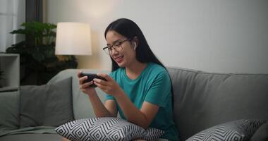 Porträt von jung asiatisch Frau tragen Brille und Kopfhörer genießt spielen online Esport Spiele auf Smartphone Sitzung auf Sofa im das Leben Zimmer beim Zuhause, Gamer Lebensstil Konzept. foto