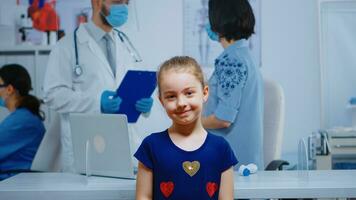 Porträt von Mädchen lächelnd im medizinisch Büro während Mutter reden mit Arzt im Hintergrund. Spezialist im Medizin mit Schutz Maske Bereitstellung Gesundheit Pflege Dienstleistungen, Beratung im Krankenhaus Klinik foto