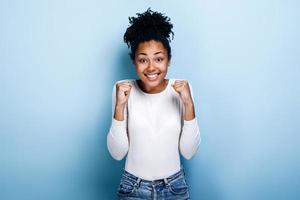überglückliches afroamerikanisches tausendjähriges Mädchen, das auf blauem Studiohintergrund isoliert ist, Triumph gute Nachrichten erhalten. Gewinnerkonzept