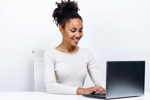 glückliche junge Frau, die am Tisch sitzt und Laptop auf weißem Hintergrund benutzt foto
