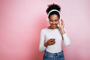 Attraktives Mädchen mit Kopfhörern auf rosafarbenem Hintergrund im Studio schaltet die Musik am Telefon ein foto