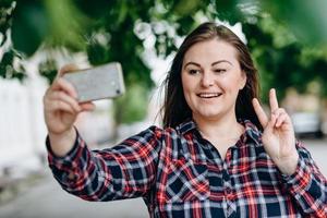 Lächelndes hübsches Mädchen, das Friedensgeste zeigt, während es ein Selfie macht, das über dem Stadthintergrund isoliert ist? foto