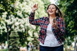 schöne Frau mit langen Haaren, in einem karierten Hemd, in Kopfhörern, mit einem Smartphone in der Hand im Freien, Spaß haben, springen foto