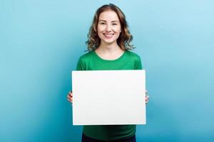 blondes Mädchen, das in grünem Pullover trägt, der einen weißen Tafelkopierraum vor blauem Hintergrund hält foto