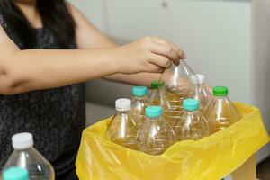 Zuhause recyceln Öko Grün Null Konzept Frau werfen leeren Plastik Flasche im Recycling Behälter mit Gelb Müll Taschen beim heim. foto