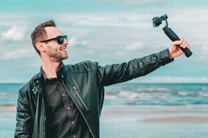 Porträt von lächelnd Blogger im Sonnenbrille Herstellung Selfie oder Streaming Video beim das Strand mit Aktion Kamera mit kardanisch Kamera Stabilisator. Mann im schwarz Kleider Herstellung Fotos