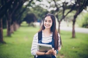Schöne asiatische Studentin, die Bücher hält und in die Kamera lächelt und im Sommer ein Lern- und Bildungskonzept im Park zum Entspannen bei Sonnenuntergang mit warmem Licht bietet