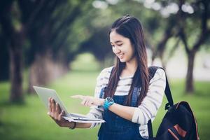 Schöne asiatische Studentin, die Bücher hält und in die Kamera lächelt und im Sommer ein Lern- und Bildungskonzept im Park zum Entspannen bei Sonnenuntergang mit warmem Licht bietet