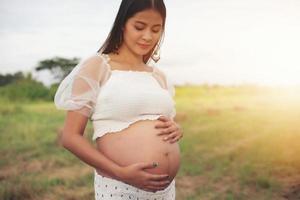 glückliche und stolze schwangere asiatische frau, die ihren bauch in einem park bei sonnenaufgang betrachtet foto