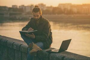 gut aussehend Geschäftsmann mit persönlich Veranstalter und Laptop während Sitzung durch das Fluss.getönt Bild foto