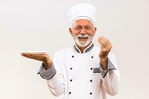 Bild von Senior Koch mit leeren Teller zeigen köstlich Zeichen auf grau Hintergrund. foto