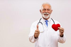 Porträt von Senior Arzt mit Stethoskop zeigen Herz gestalten und Daumen oben auf grau Hintergrund. foto