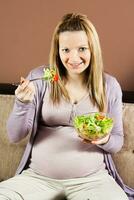 schwanger Frau Sitzung auf das Sofa und Essen Salat foto