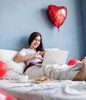 Junge glückliche brünette Frau, die im Bett mit roten herzförmigen Ballons sitzt und mit ihrem Freund auf dem Laptop plaudert und Herzgeste mit den Händen zeigt
