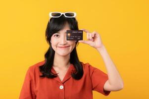 Porträt jung schön asiatisch Frau glücklich Lächeln gekleidet im Orange Kleider zeigen Anerkennung Karte isoliert auf Gelb Hintergrund. Zahlen und Kauf Einkaufen Zahlung Konzept. foto