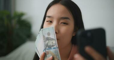 Porträt von glücklich jung asiatisch Frau Sitzung im ein aufgeregt Zustand, halten ein online Smartphone und Dollar Rechnungen im ihr Hand, spart Geld foto