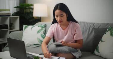 Porträt von jung asiatisch Frau prüft ihr Finanzen mit ein Taschenrechner auf Smartphone und macht Anmerkungen im ein Notizbuch während Sitzung auf das Sofa beim Zu Hause zu Hause Finanzen foto