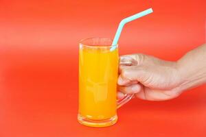 schließen oben von Mann Hand halten Glas von Orange Saft mit Trinken Stroh, auf rot Hintergrund. Konzept, Morgen erfrischend Getränk. süss, gereizt und hoch Vitamin c. foto