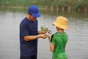 asiatisch Vater und Sohn ist erkunden Ökologie im See, Vater halt Glas Krug von Wasser, Sohn hält Vergrößerung Glas. Konzept, Studie Ökologie Gegenstand erkunden Wasser von Natur Quelle.Bildung, Leben Erfahrung foto