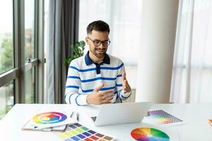 heiter Mann Designer haben Video Konferenz mit Kunden, Sitzung beim Schreibtisch im Vorderseite von Computer, halten Farbe Paletten, gestikulieren und lächelnd, Kopieren Raum foto