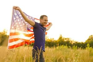 vierte von Juli. patriotisch Mann mit das National amerikanisch Flagge im das Feld. jung Mann stolz winken ein amerikanisch Flagge. Unabhängigkeit Tag. foto
