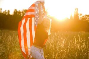 schön jung Mädchen halten ein amerikanisch Flagge im das Wind im ein Feld von Roggen. Sommer- Landschaft gegen das Blau Himmel. horizontal Orientierung. foto