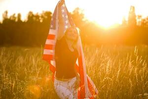 schön jung Mädchen halten ein amerikanisch Flagge im das Wind im ein Feld von Roggen. Sommer- Landschaft gegen das Blau Himmel. horizontal Orientierung. foto