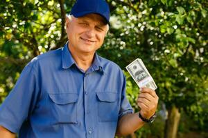 heiter Senior halten Geld Bündel im das Garten Hintergrund foto