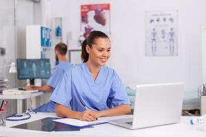 medizinisch Praktiker Sitzung beim Schreibtisch im Krankenhaus Büro mit Laptop mit Arzt im das Hintergrund. Gesundheit Pflege Arzt mit Computer im modern Klinik suchen beim Monitor, Medizin, Beruf. foto