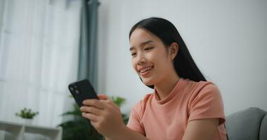 Porträt von glücklich jung asiatisch Frau entspannen auf komfortabel Couch beim Zuhause Einkaufen online auf Smartphone, lächelnd Mädchen verwenden Zelle Telefon chatten, Durchsuche kabellos Internet auf Gadget foto